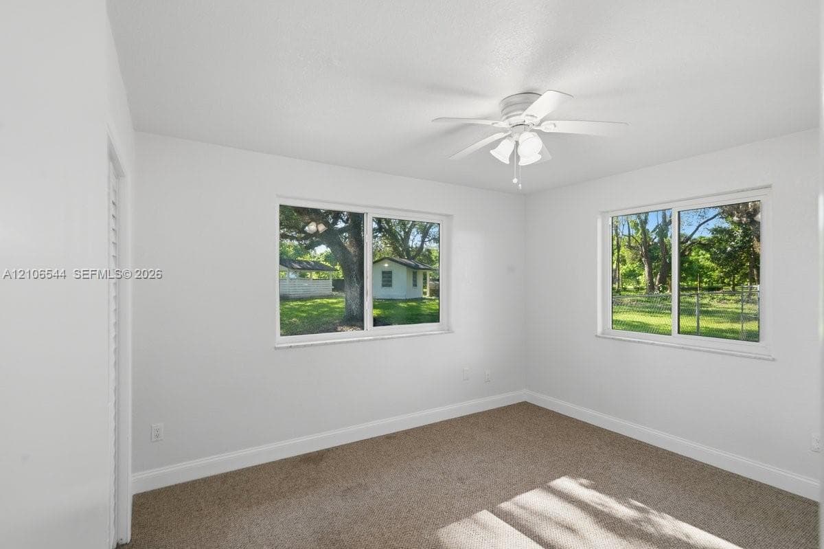 Neutral-toned interior space featuring wall-to-wall carpeting, white walls, dual windows, and a ceiling-mounted light fixture with a multi-blade fan