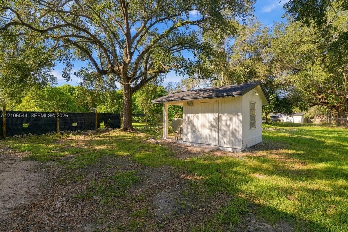 Detached storage structure featuring white siding, a shingled roof, a covered entry, and mature trees situated on a cleared lot with black perimeter fencing