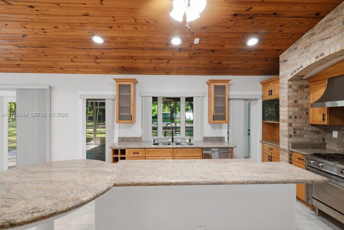 Kitchen featuring wood-plank cathedral ceilings, stone-surfaced kitchen island, custom cabinetry with glass-front uppers, brick-surround stove alcove, and stainless steel appliances