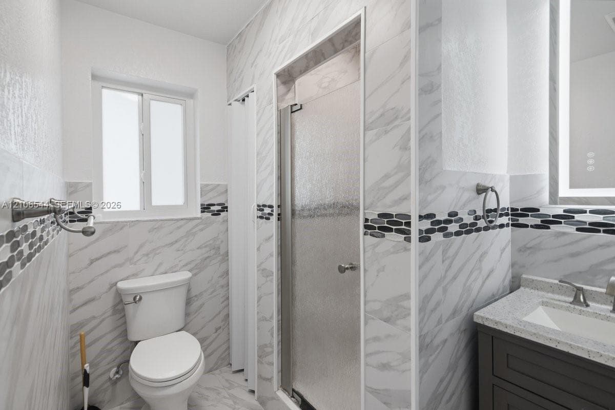 Bathroom featuring stone-finish wall tile with horizontal decorative accent inlay, enclosed shower with textured glass door, dark cabinetry with light-toned countertop, and integrated-light vanity mirror