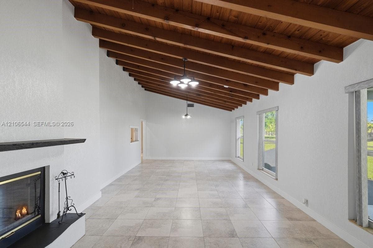 Vaulted ceiling featuring exposed wood beams, neutral tile flooring, fireplace with a matte finish mantel, and large windows