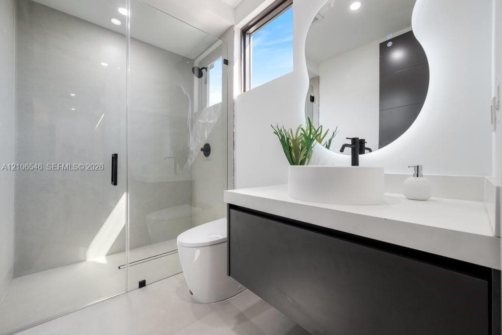 Floating dark-finish vanity with a white countertop and vessel sink, frameless glass walk-in shower with matte black fixtures, and a wall-mounted back-lit organic-shaped mirror