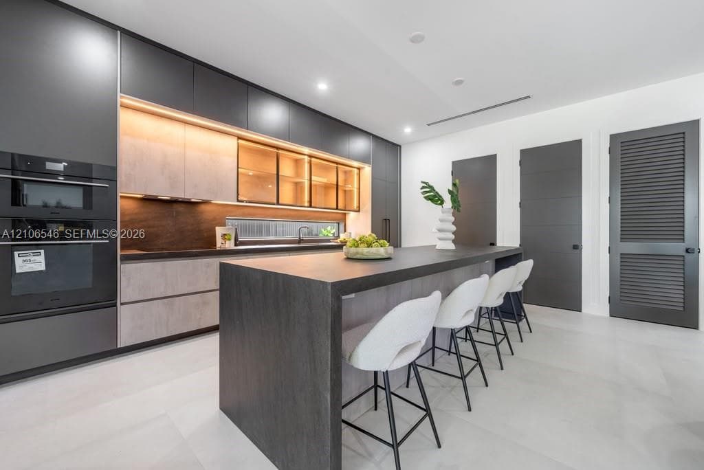 Modern kitchen featuring floor-to-ceiling dark cabinetry, a large kitchen island with a dark countertop, integrated double wall ovens, wood-finish lower cabinetry, and illuminated glass-front upper cabinets