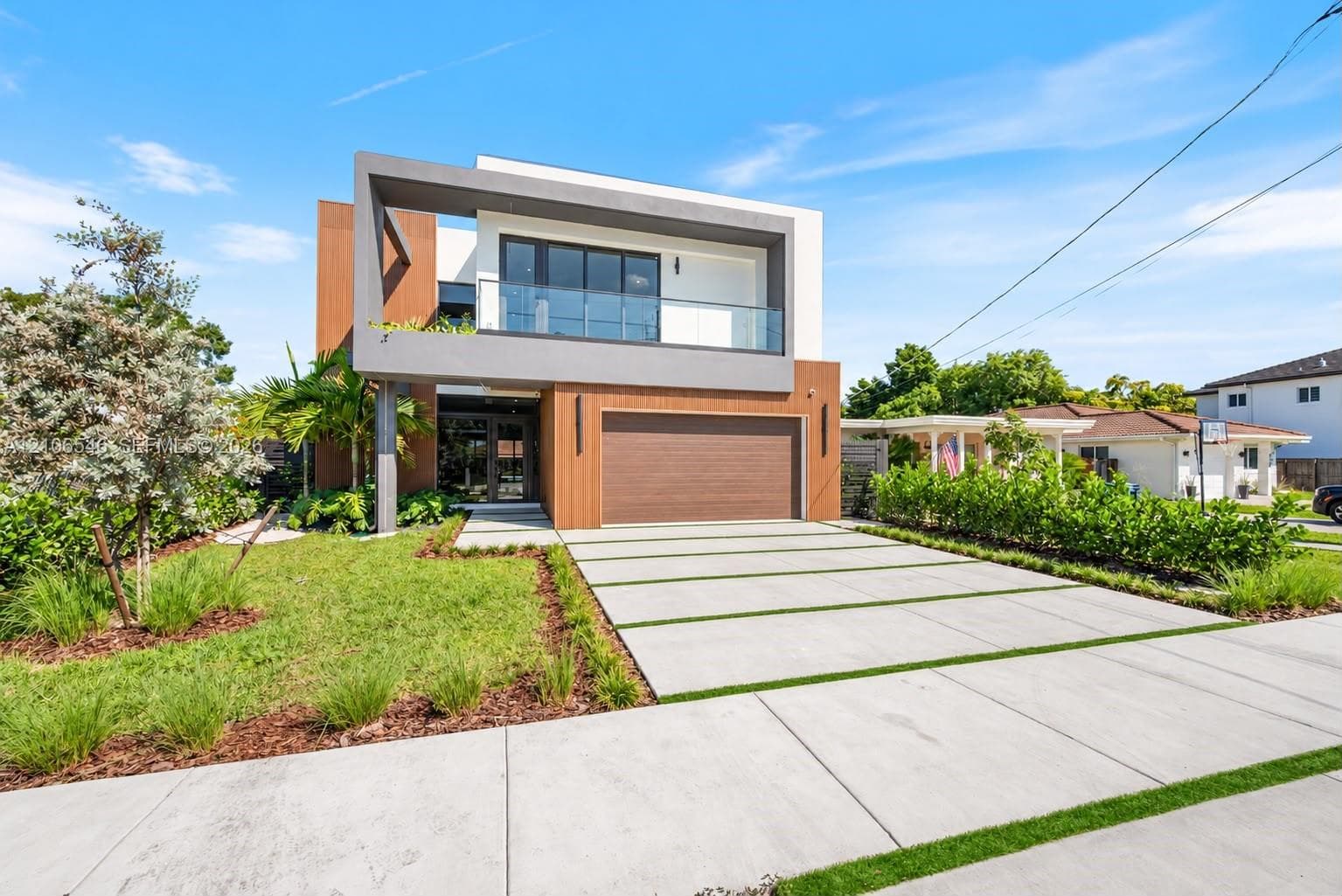 Modern architectural exterior featuring wood-tone siding, a concrete paver driveway with turf accents, a glass-railed second-story balcony, and a two-car garage