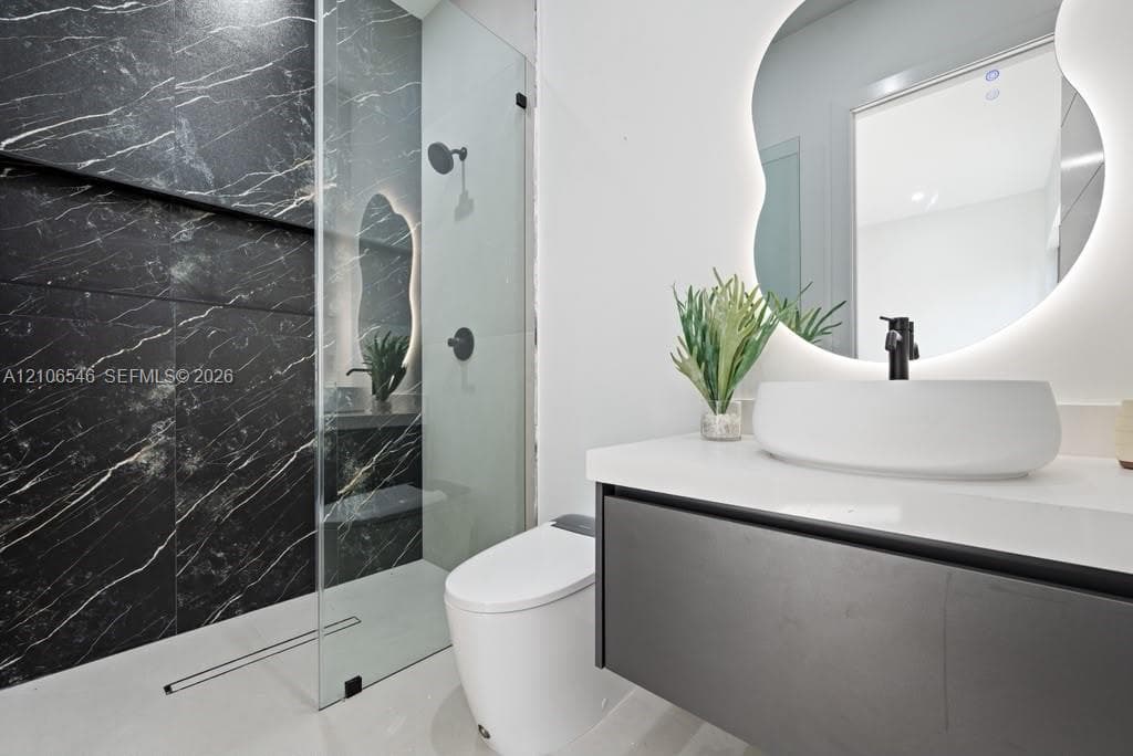 Floating vanity with white countertop and vessel sink, frameless glass shower enclosure, floor-to-ceiling dark tile shower surround, matte black hardware, and backlit organic-shaped mirror