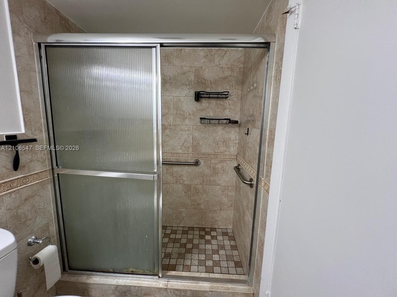 Walk-in shower enclosure featuring ribbed sliding glass doors, neutral-toned tiled walls with decorative border accents, mosaic tile flooring, and integrated metal grab bars