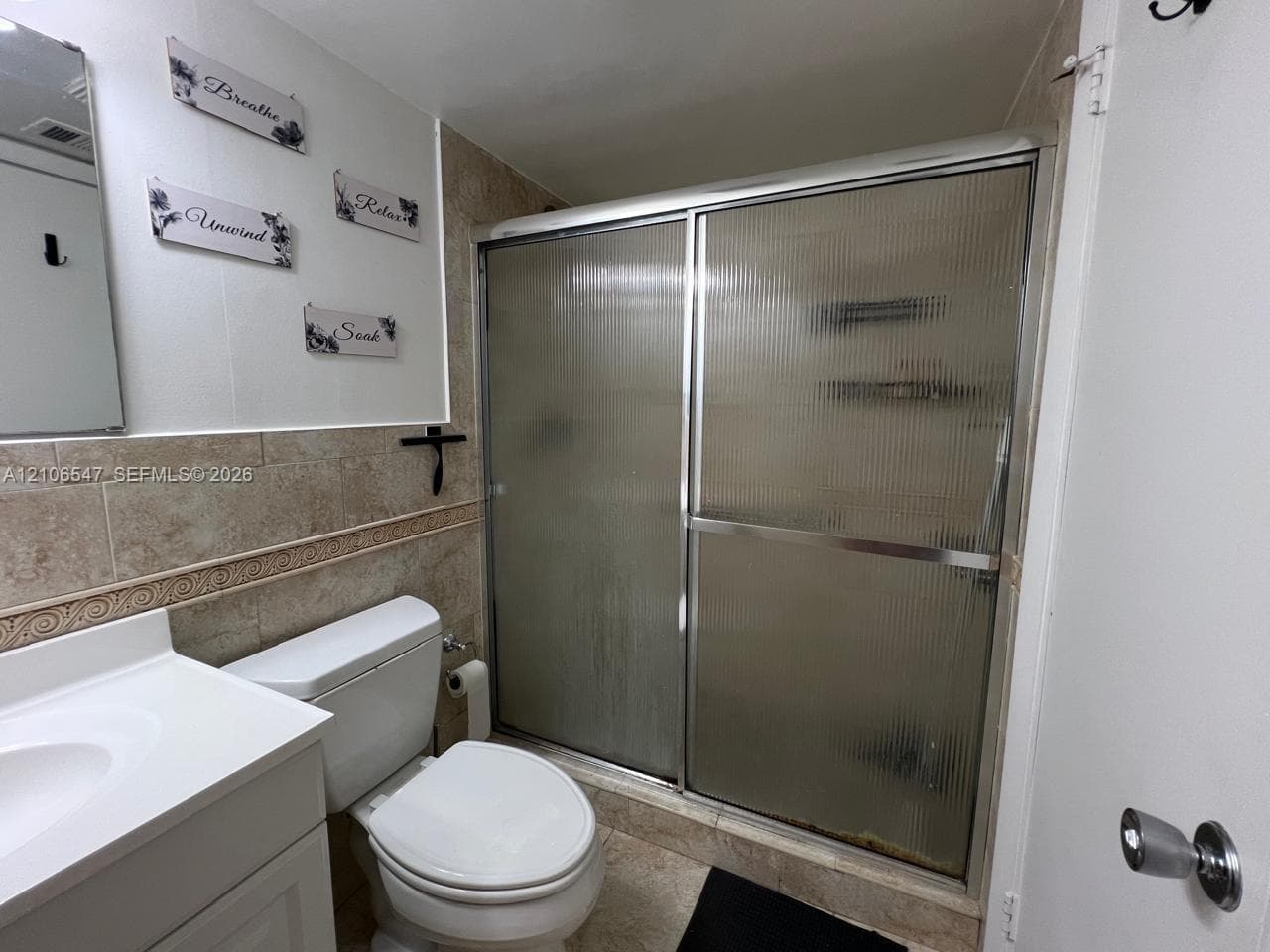 Bathroom featuring a white vanity with an integrated basin, a standard toilet, and a walk-in shower enclosed by reeded glass sliding doors with metallic framing, finished with wall-to-wall ceramic tile surround and coordinating floor tile