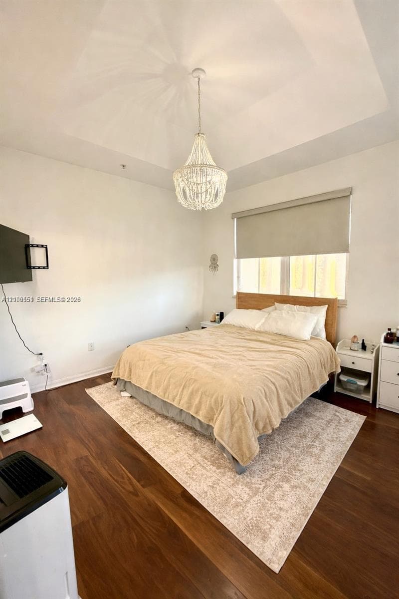 Tray ceiling featuring a decorative beaded chandelier, dark wood-finish flooring, and window with a neutral roller shade