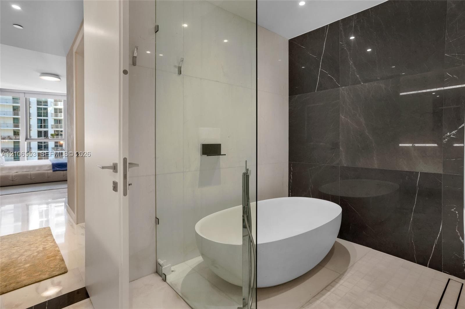 Freestanding oval soaking tub enclosed in a glass shower surround, dark stone-finish wall cladding, recessed ceiling lighting, and polished stone-finish flooring