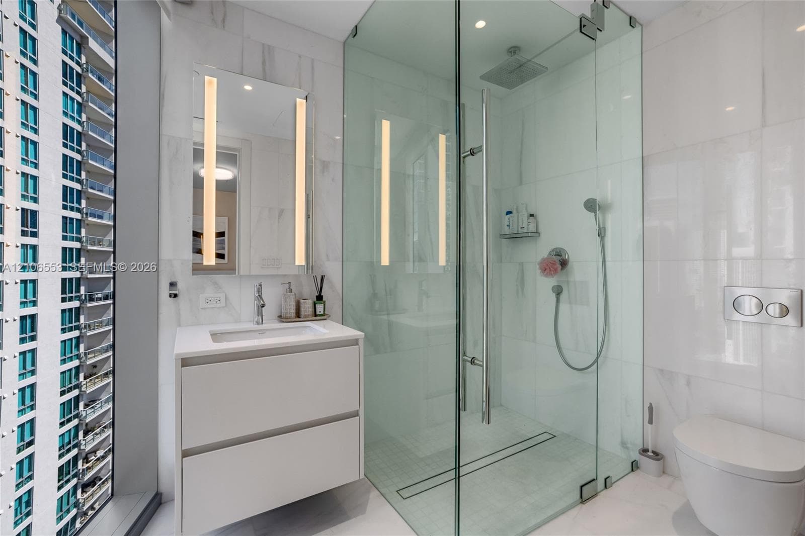 Bathroom featuring a floating vanity with a rectangular sink, wall-mounted mirror with integrated lighting, glass-enclosed shower with rainfall head and handheld sprayer, wall-mounted toilet, and floor-to-ceiling tile finishes