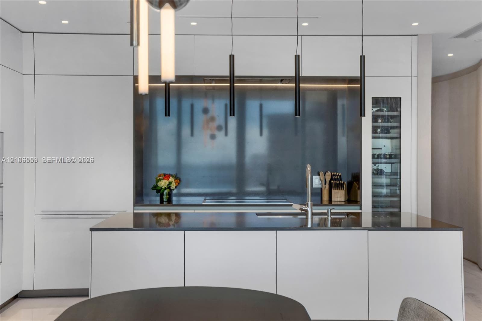 Minimalist kitchen featuring flat-panel cabinetry, a stainless steel backsplash, an integrated wine refrigerator, linear pendant lighting, and a solid-surface island with a built-in sink