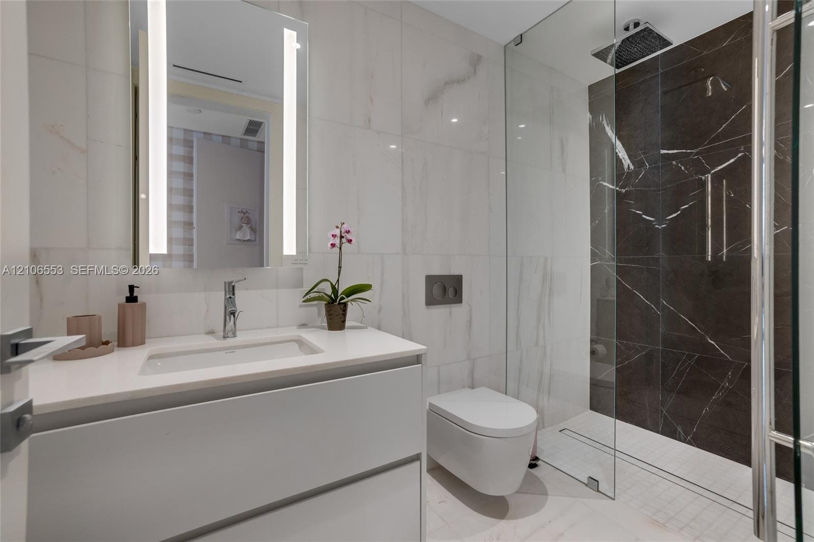 Floating vanity with a quartz countertop, integrated rectangular sink, wall-mounted dual-flush toilet, glass-enclosed shower with dark stone-finish tiling, and a backlit vanity mirror