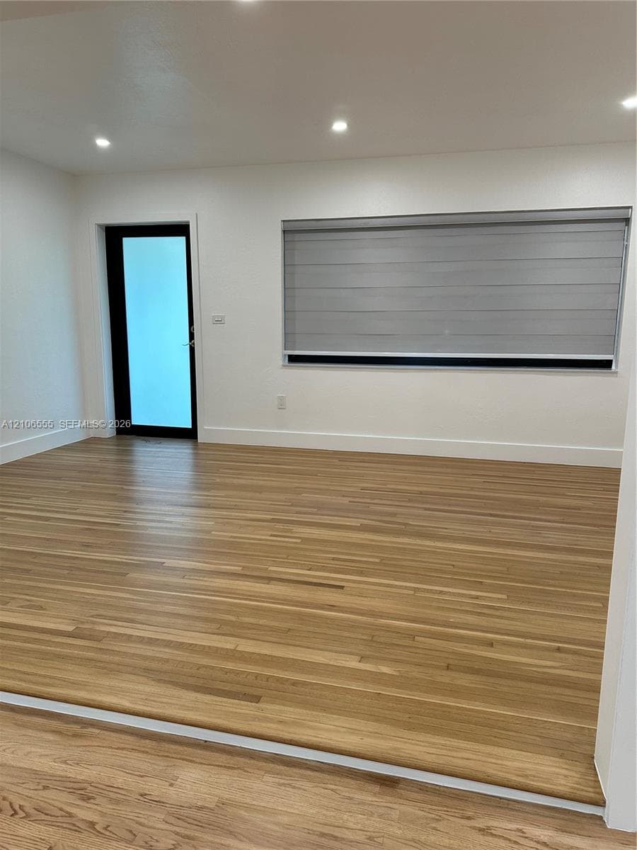 Neutral-toned interior featuring wood-finish flooring, recessed lighting, a glass-panel entry door with a black frame, and a wide window with integrated horizontal blinds