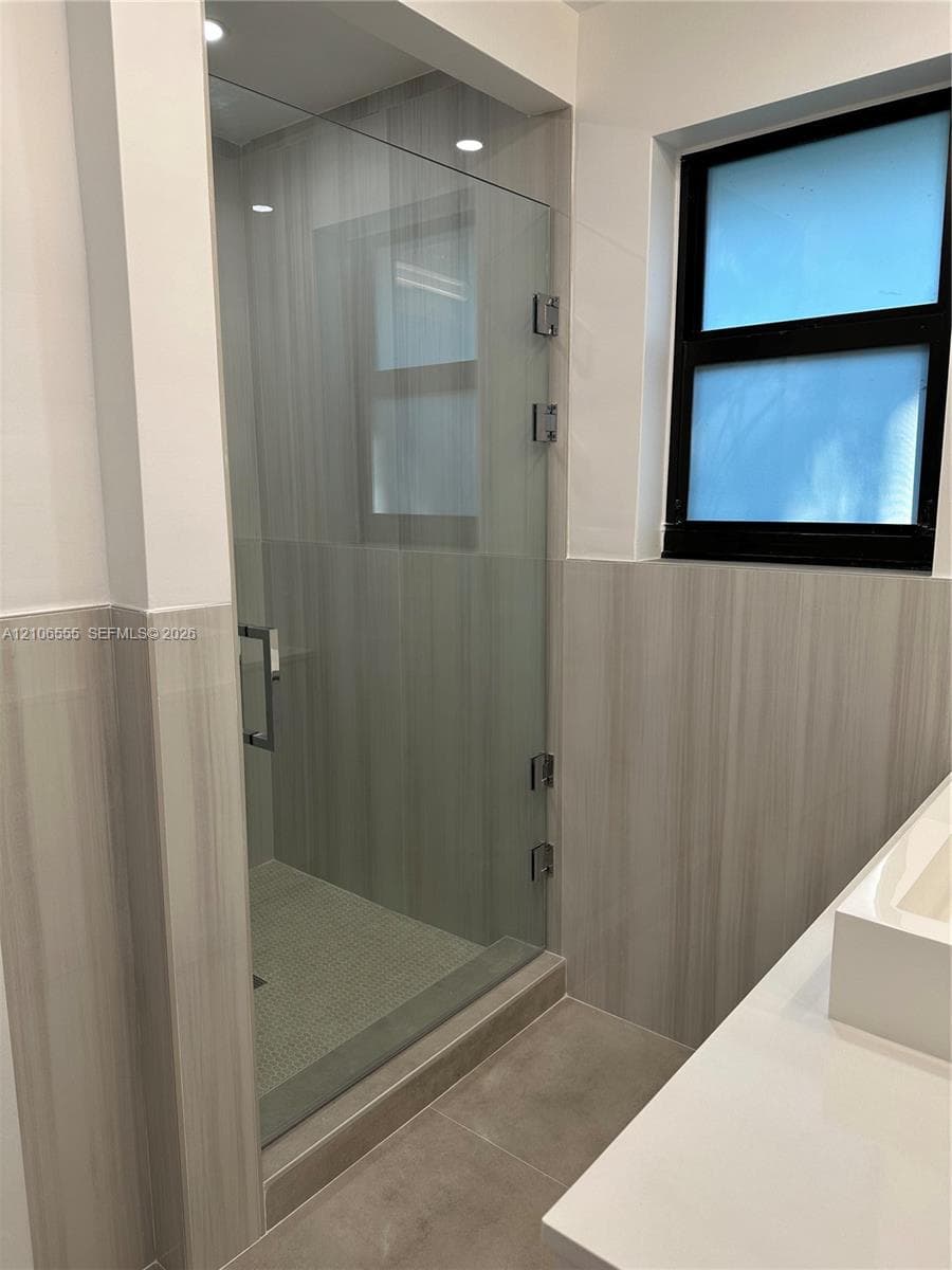 Walk-in shower featuring a frameless glass door, stone-finish tile surround, integrated pebble-style floor tiling, solid surface vanity countertop, and a window with a black frame