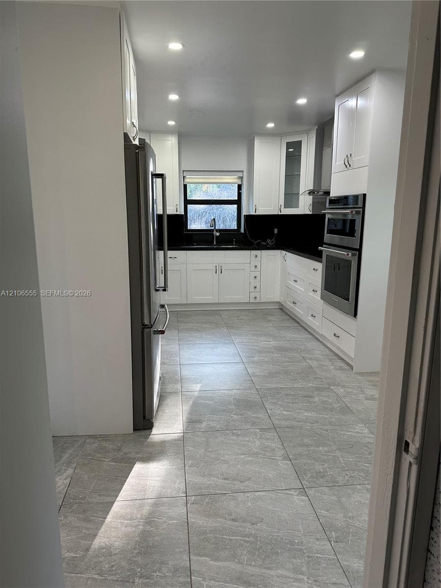 Kitchen featuring white shaker-style cabinetry, stainless steel appliances, dark stone-finish countertops, recessed lighting, and large-format tile flooring