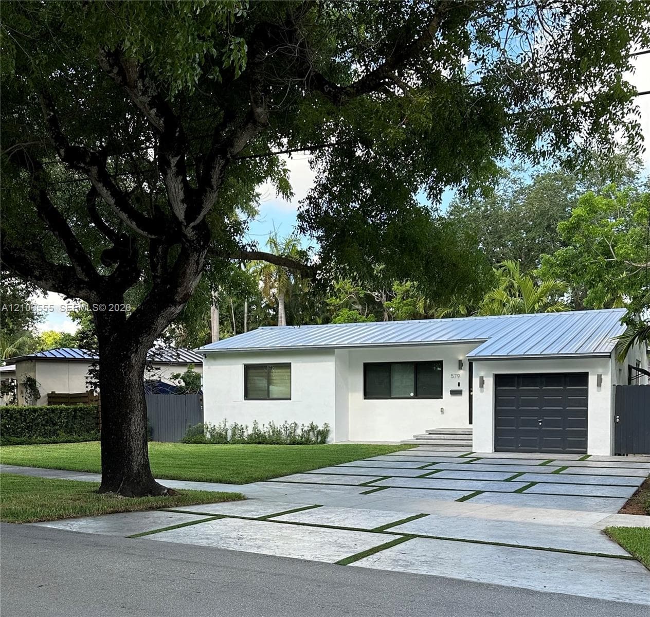 Single-story exterior featuring a white stucco facade, metal roof, paver driveway with turf accents, attached garage, and mature landscaping