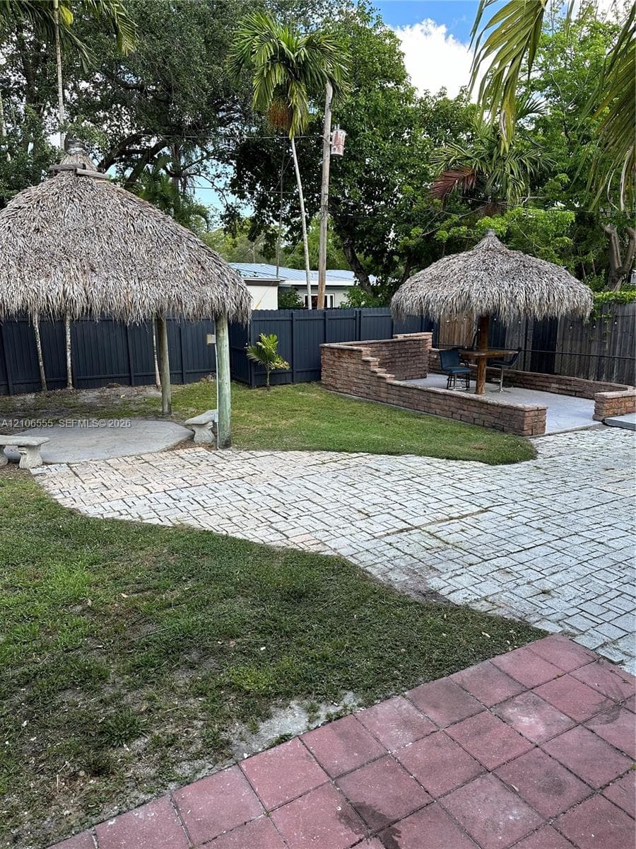 Backyard featuring dual thatched-roof structures, tiered masonry walls, patterned paver hardscaping, and privacy fencing