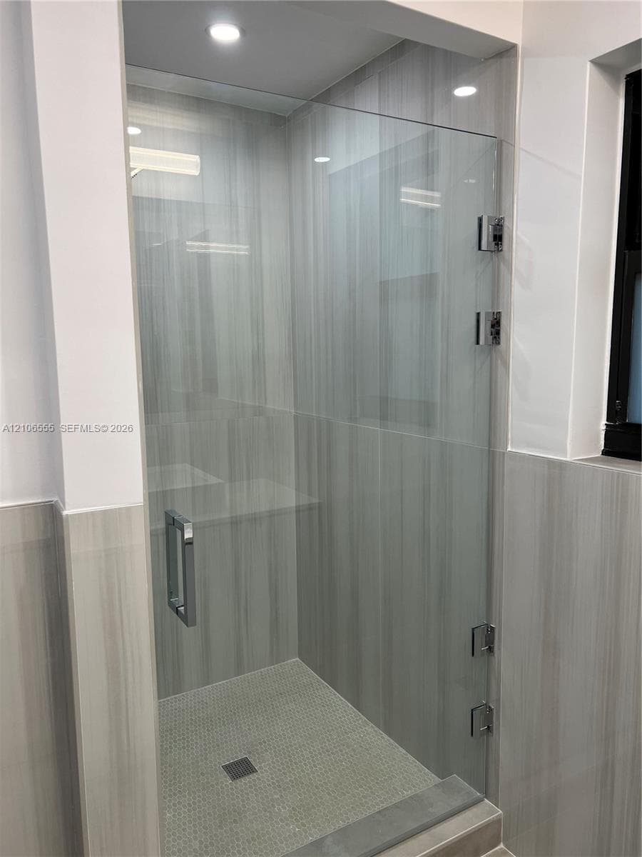 Walk-in shower featuring large-format vertical-pattern wall tile, frameless glass door with polished chrome hardware, stone-finish floor tiling, and integrated recessed lighting