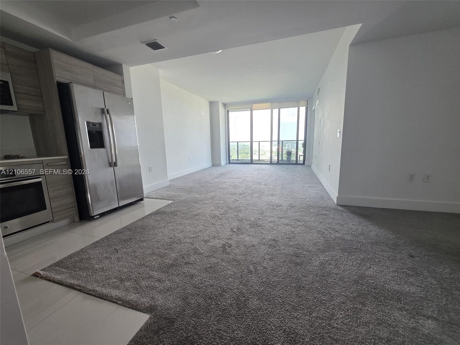 Kitchen featuring textured cabinetry, stainless steel appliances, and tile flooring, opening to a spacious area with carpeted flooring and sliding glass balcony doors