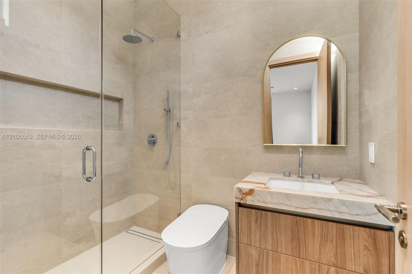 Bathroom featuring floor-to-ceiling textured tile, wood-finish vanity with stone countertop, frameless glass shower enclosure with rainfall showerhead, and arched wall-mounted mirror
