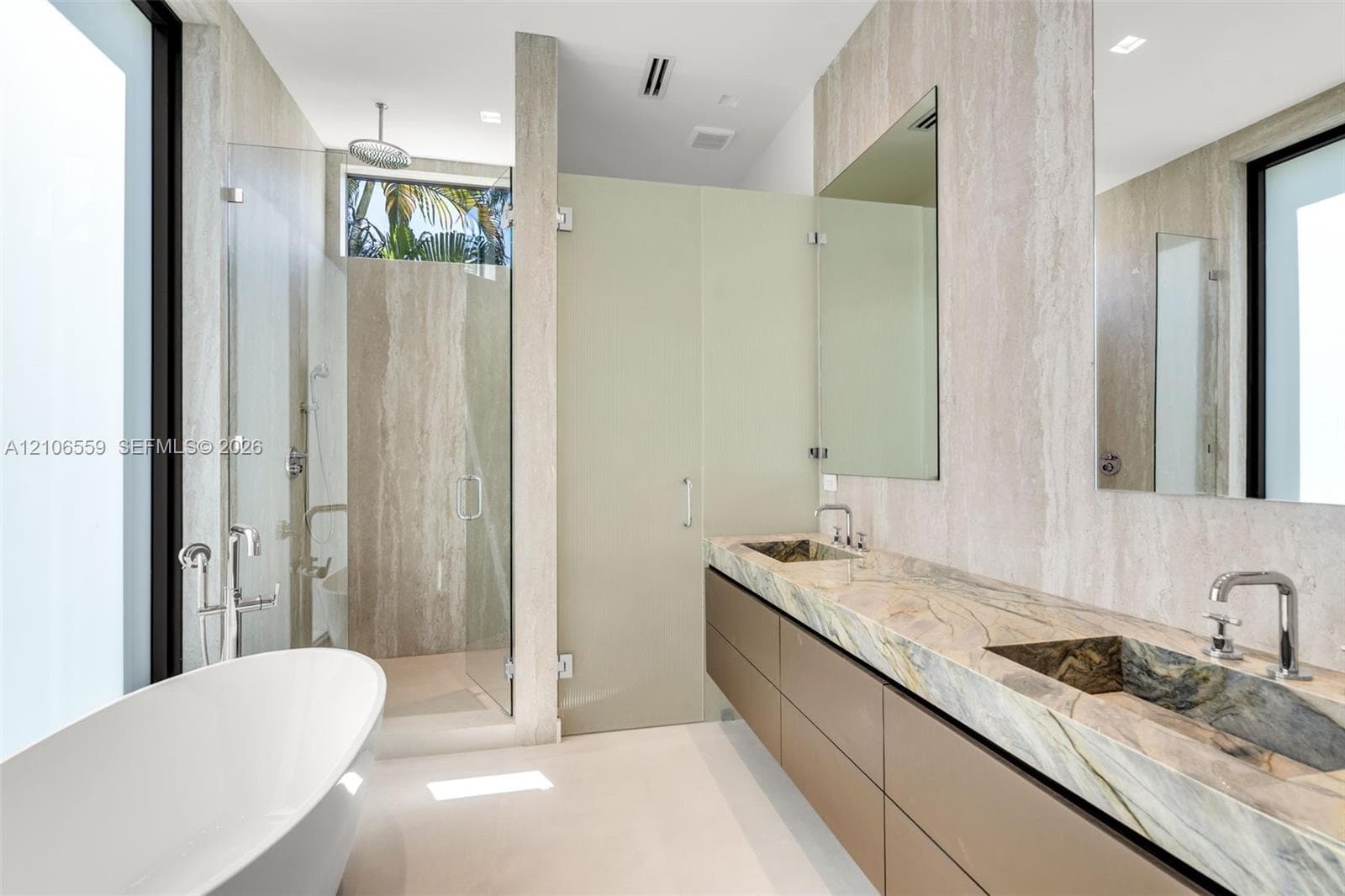 Floating dual-sink vanity featuring integrated stone sinks, natural stone slab countertop, and matte metallic fixtures, paired with a freestanding soaking tub, glass-enclosed shower, and floor-to-ceiling natural stone wall finishes