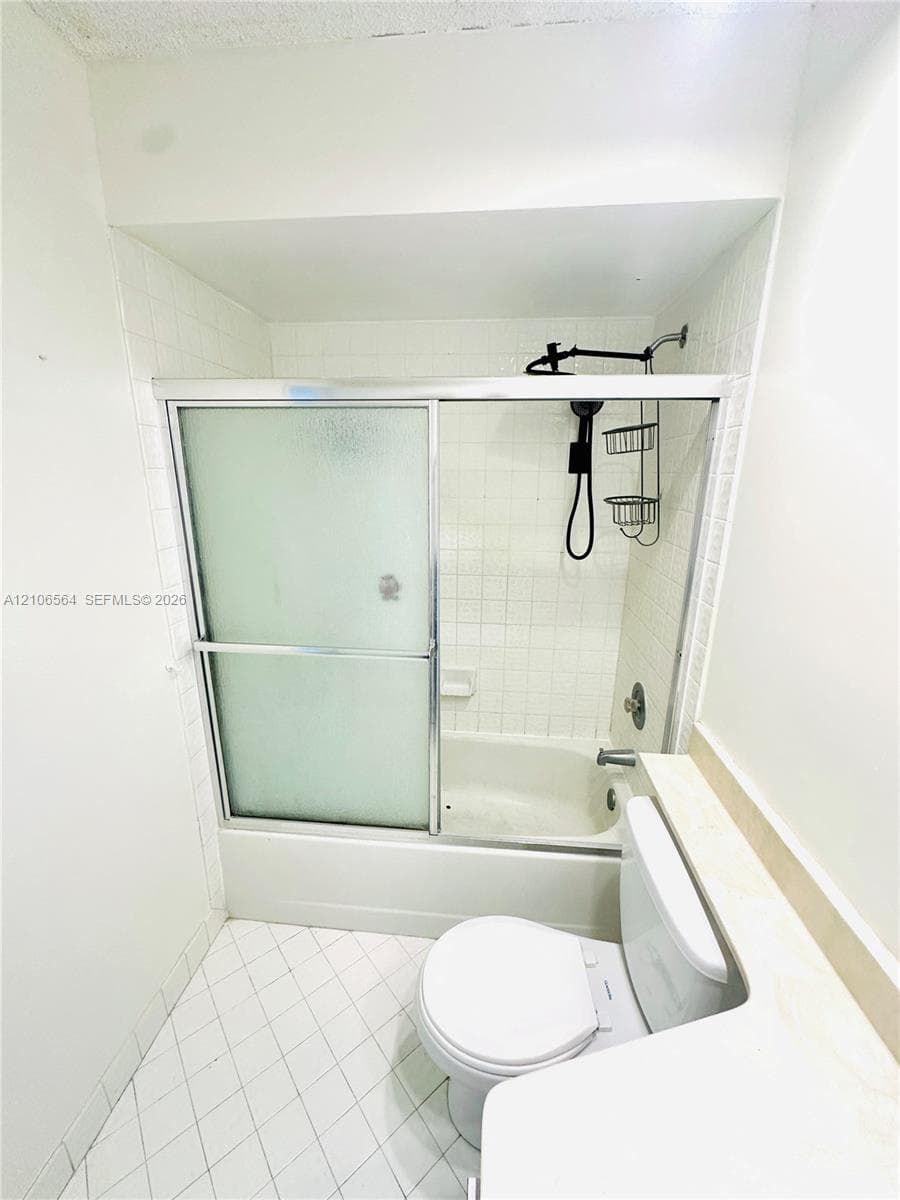 Full bathtub featuring a tiled surround, sliding frosted glass enclosure, and a wall-mounted shower fixture with a handheld attachment, complemented by white diamond-patterned flooring