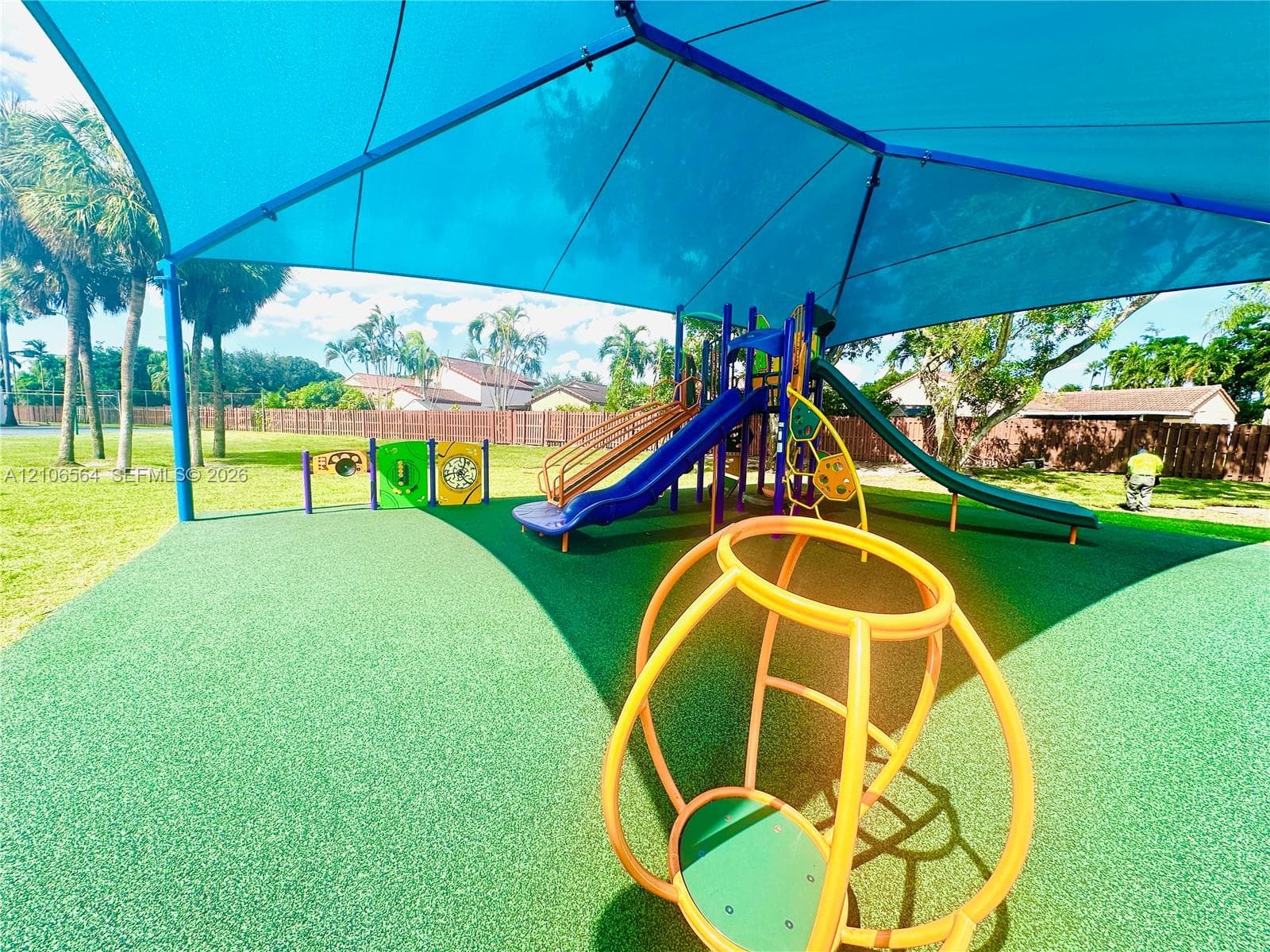 Outdoor recreational area featuring a large blue shade canopy, multi-level playground structure, spiral and straight slides, and durable rubberized ground surfacing