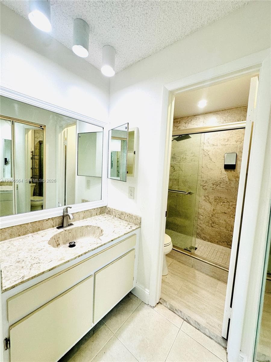 Vanity featuring a stone-composite countertop with an integrated basin, large wall-mounted mirror, overhead cylindrical track lighting, and a glass-enclosed shower with tile surround