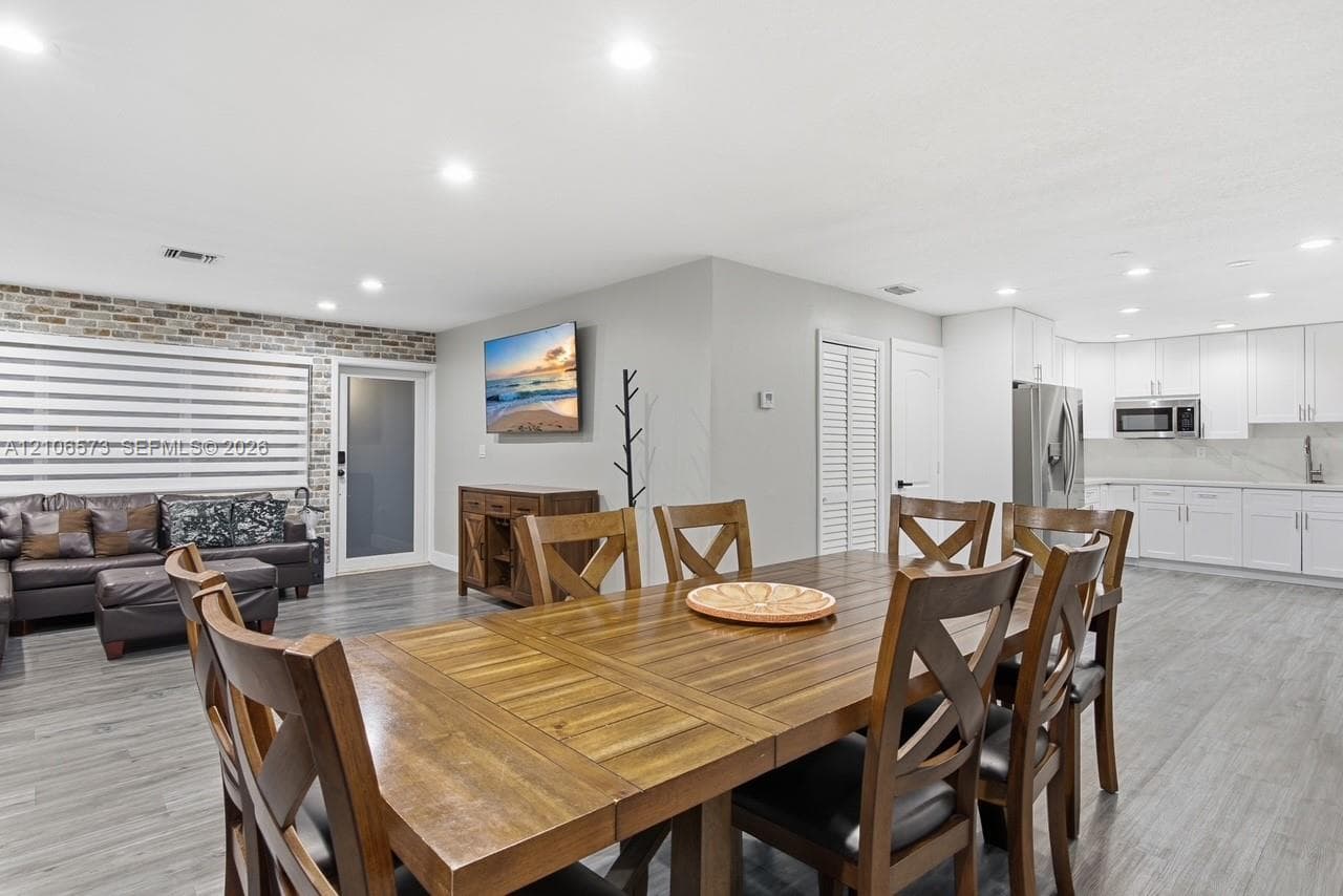 Open-concept living space featuring wood-finish flooring, recessed lighting, exposed brick accent wall, and a kitchen with white cabinetry and stainless steel appliances
