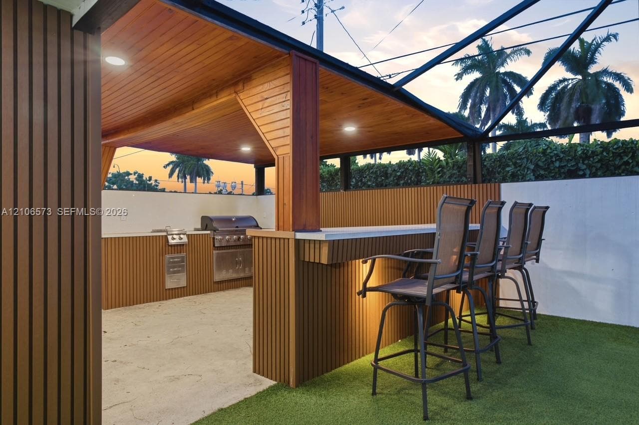 Covered outdoor kitchen featuring vertical slat siding, recessed ceiling lighting, a stone-finish bar counter, and built-in stainless steel appliances