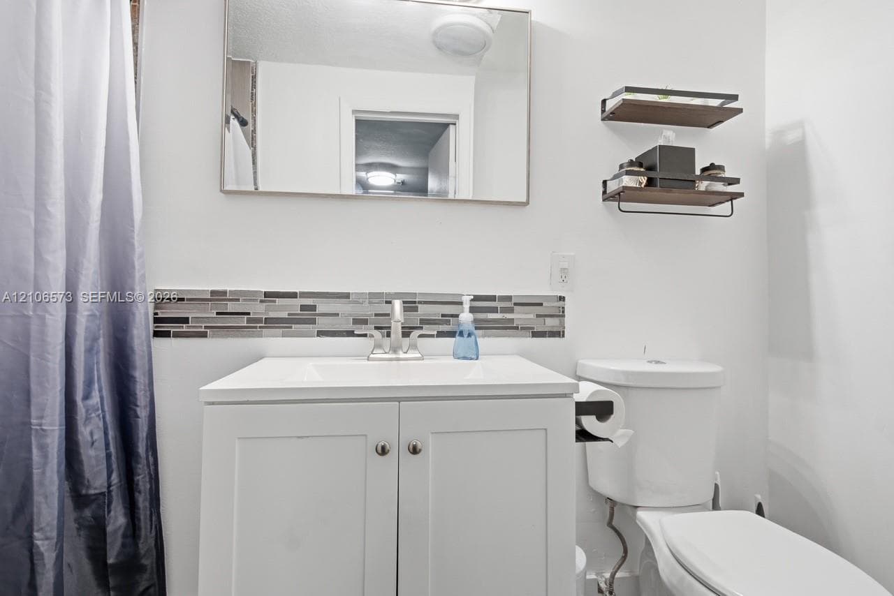 Bathroom featuring a white vanity cabinet with a integrated sink, metallic faucet, rectangular wall-mounted mirror, decorative horizontal mosaic tile backsplash, and two tiered wall-mounted storage shelves