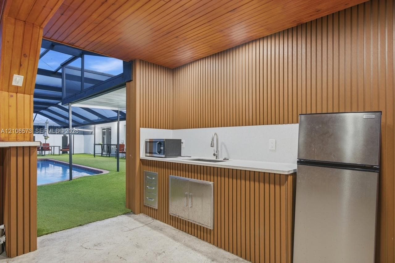 Covered outdoor kitchen featuring vertical wood-paneled walls and ceiling, stainless steel refrigerator, undermount sink with gooseneck faucet, and built-in stainless steel cabinetry