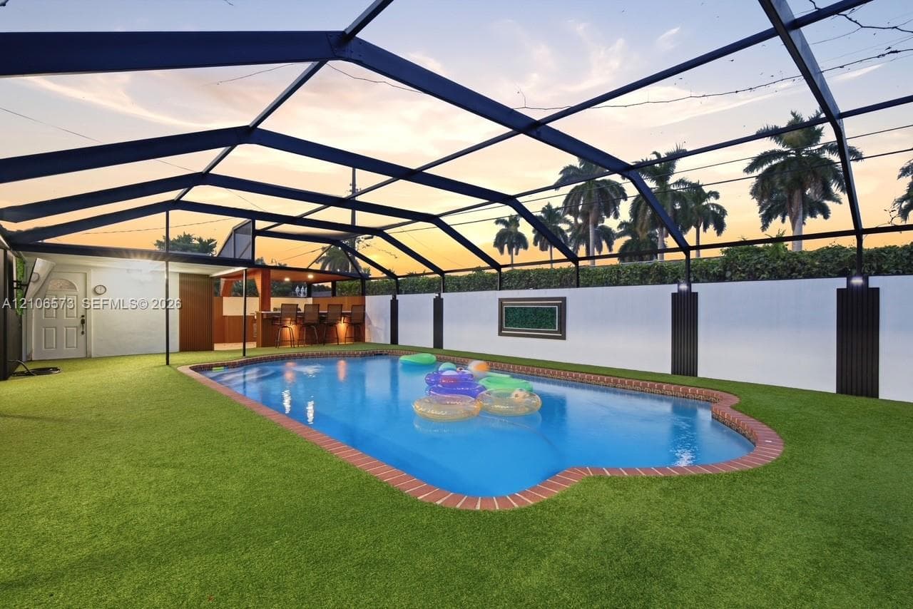 In-ground swimming pool with brick paver coping, fully enclosed by a screened lanai structure, featuring a built-in outdoor bar and synthetic turf surrounding