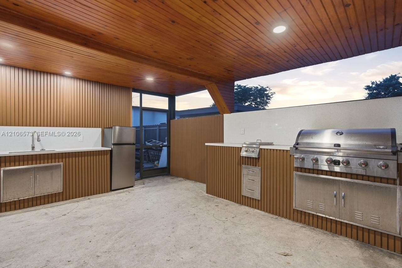 Covered outdoor kitchen featuring vertical wood-slat cabinetry, expansive stone countertops, stainless steel built-in grill, integrated sink, and recessed lighting