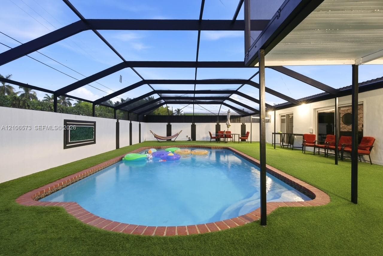 In-ground pool with brick coping, enclosed by a screened lanai structure, featuring artificial turf surfacing and a perimeter privacy fence
