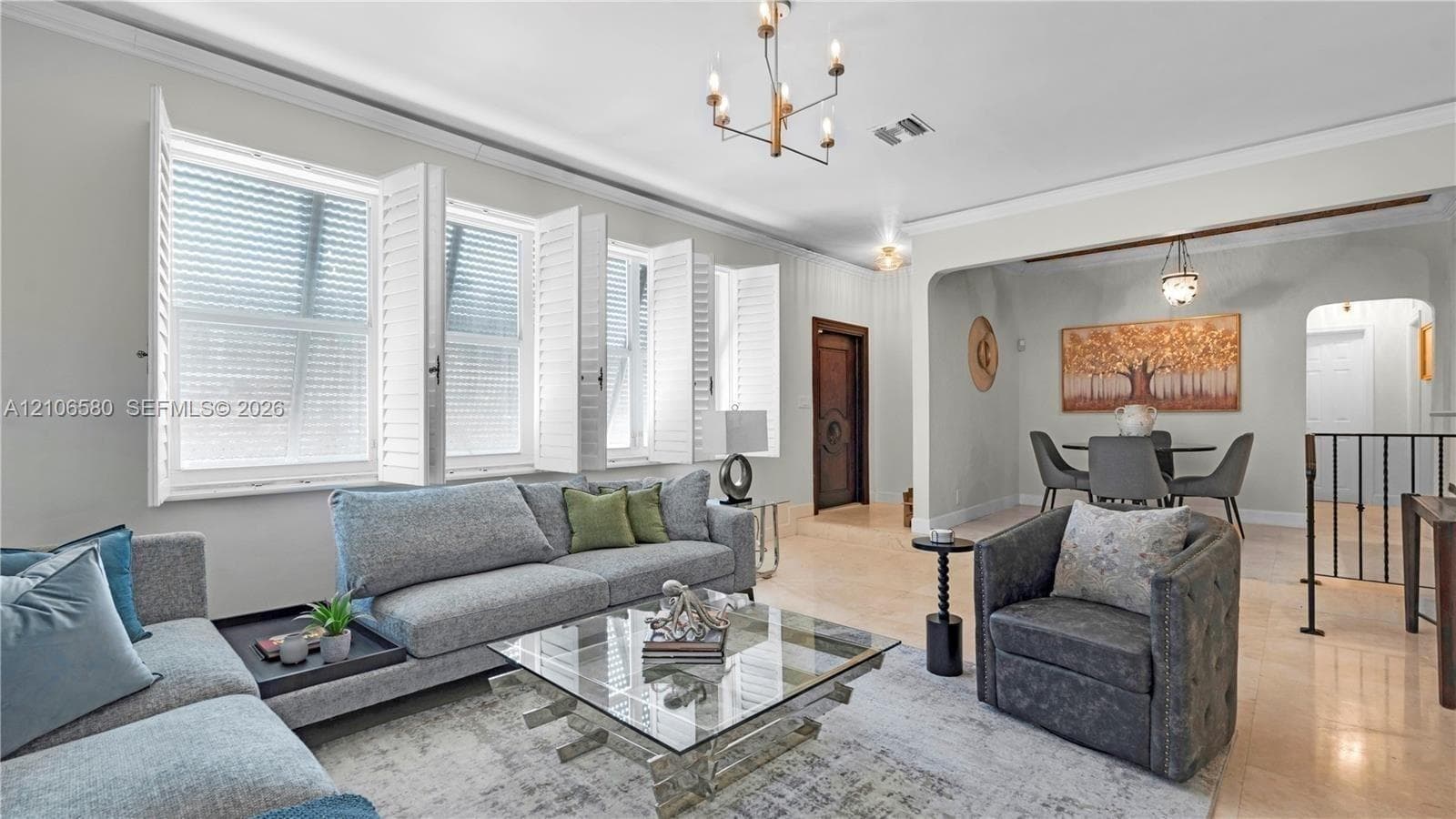 Living area featuring tile flooring, crown molding, multi-pane windows with plantation shutters, and a brass-finish chandelier
