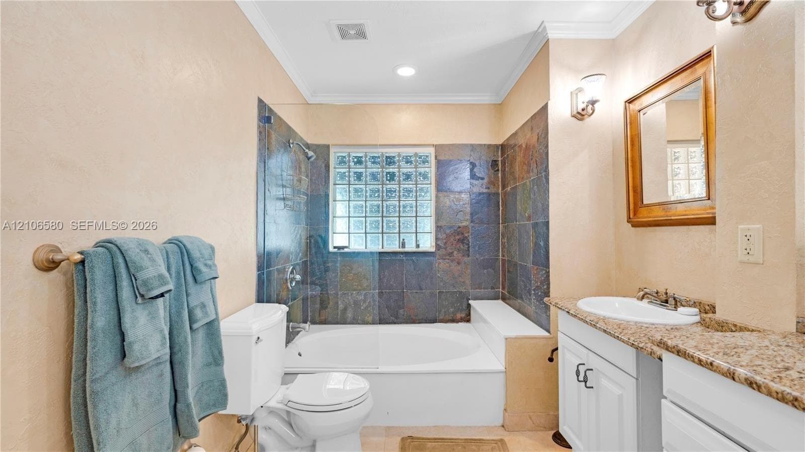 Bathroom featuring a stone-tile tub surround, glass-block window, undermount sink with a stone countertop, white shaker-style vanity cabinetry, and wall-mounted light fixtures