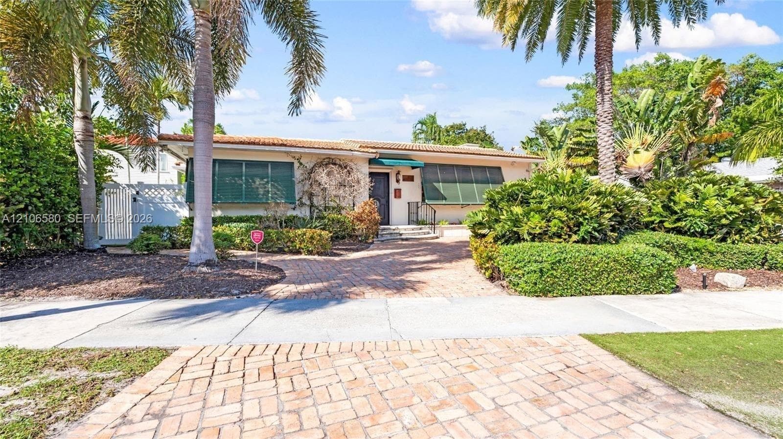 Single-story residence featuring a brick-paved driveway, clay tile roof, metal window awnings, and mature landscaping