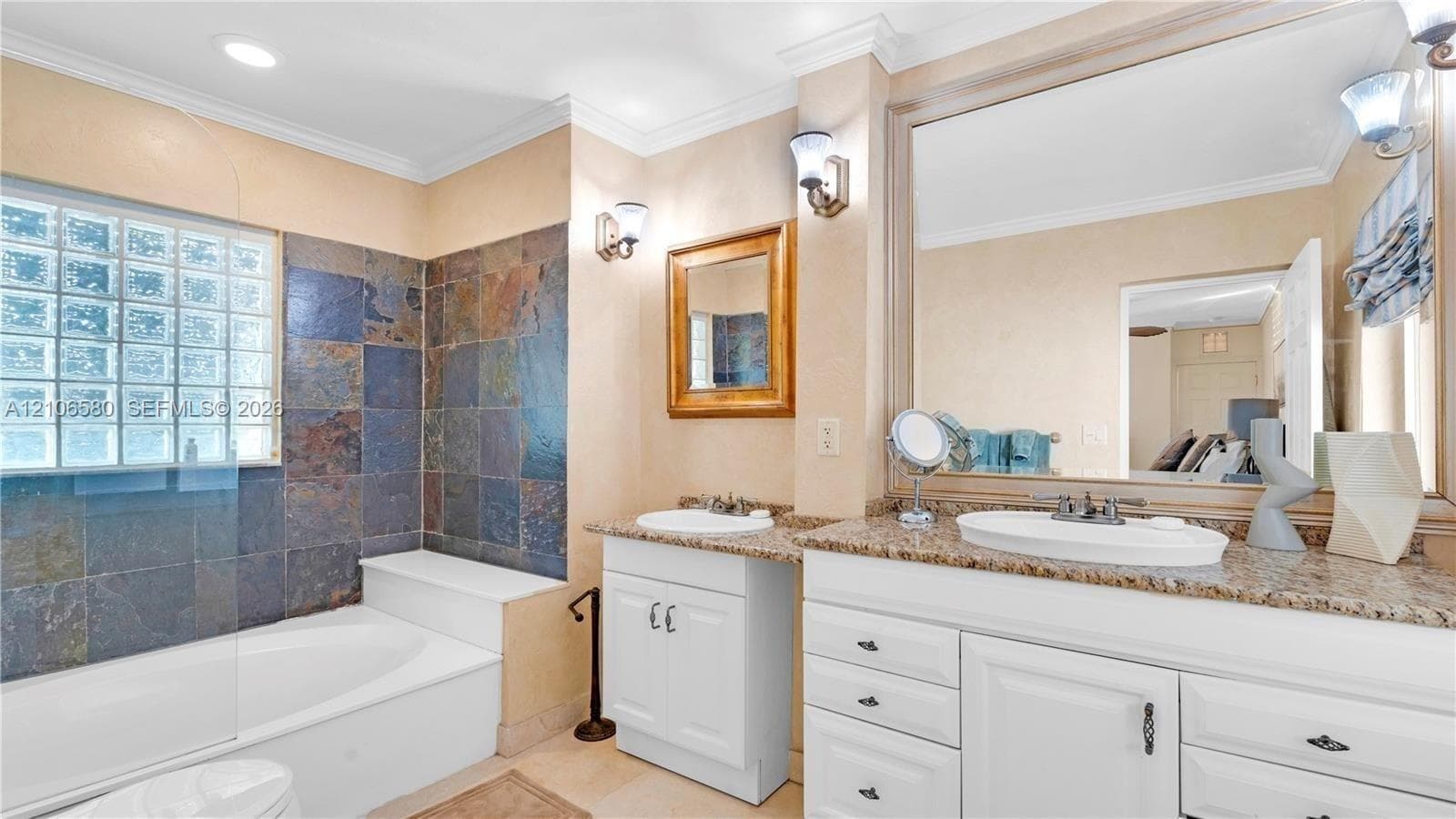Bathroom featuring dual white vanities with stone countertops, undermount sinks, glass block window, and slate tile surround at the soaking tub