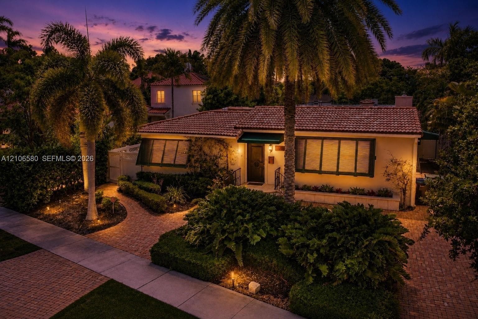 Single-story residence featuring a tile roof, paved walkway, mature palm trees, manicured hedges, and exterior wall-mounted lighting