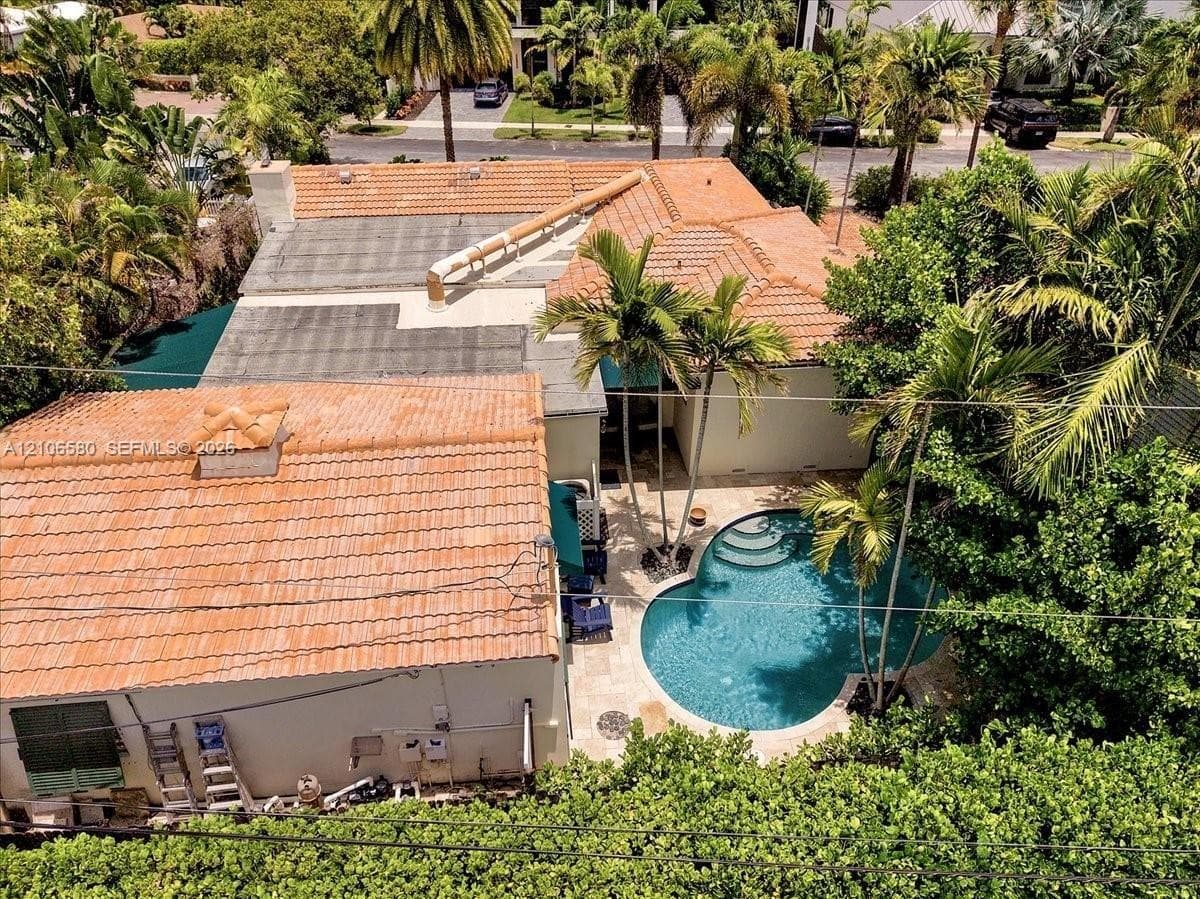 Aerial perspective of a property featuring a multi-section clay tile roof, a curved swimming pool, stone paver decking, and mature palm trees