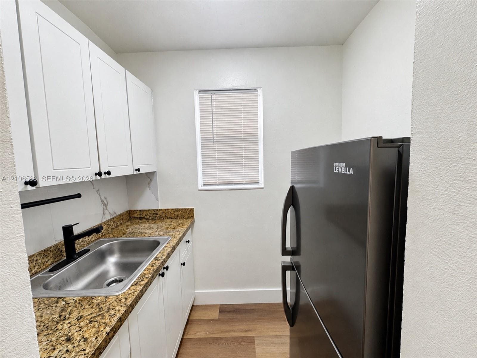 Kitchen featuring white shaker-style cabinetry, speckled stone countertops, a stainless steel sink with a matte black faucet, and wood-finish flooring