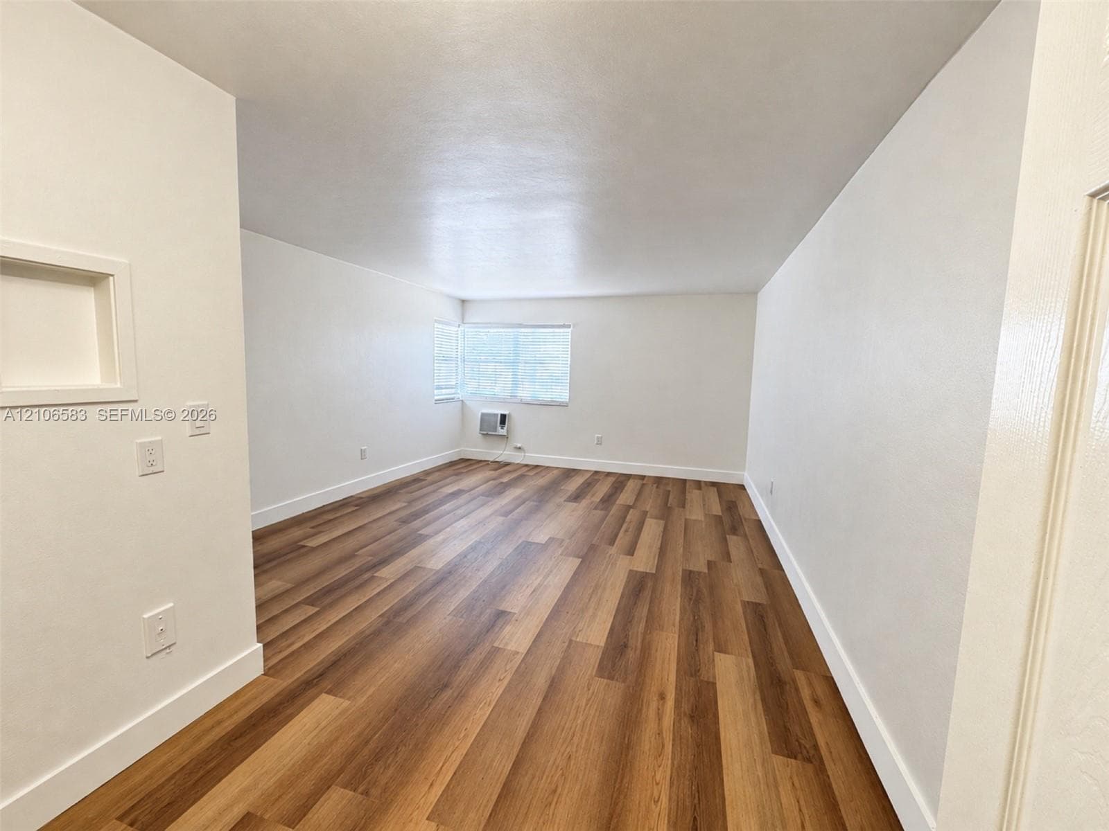 Interior space featuring wood-finish flooring, white walls, a recessed wall niche, and a window with a wall-mounted air conditioning unit