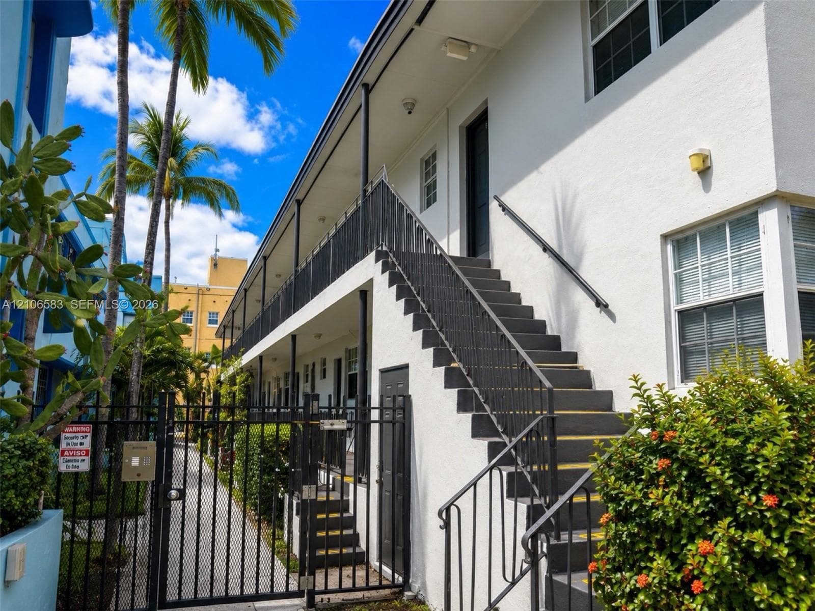 Exterior facade featuring white stucco finish, multi-level metal staircase with black railing, grid-patterned windows, and a secure metal perimeter gate