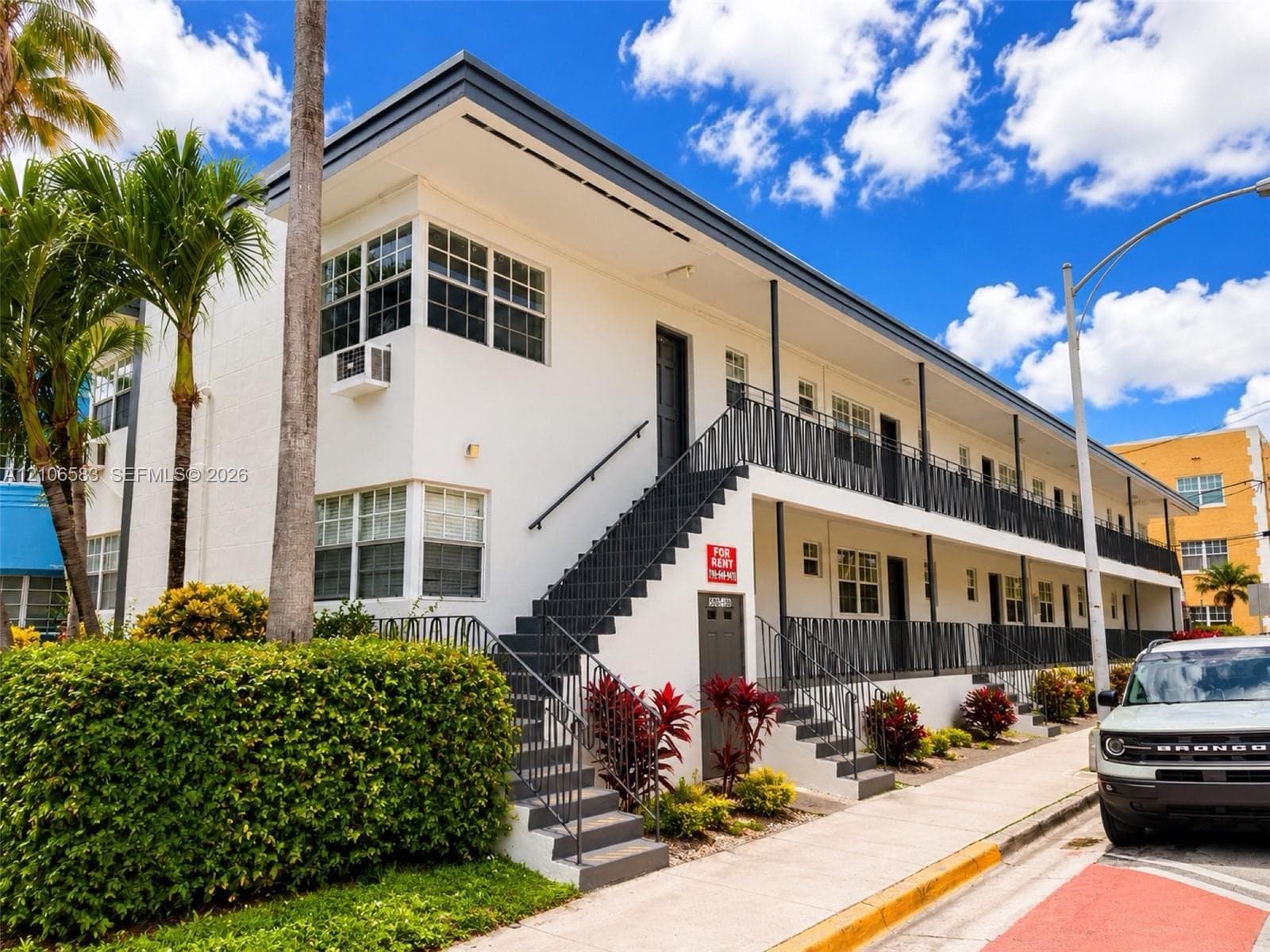 Two-story mid-century modern exterior featuring a smooth stucco finish, metal railings, exterior staircase, and mature tropical landscaping