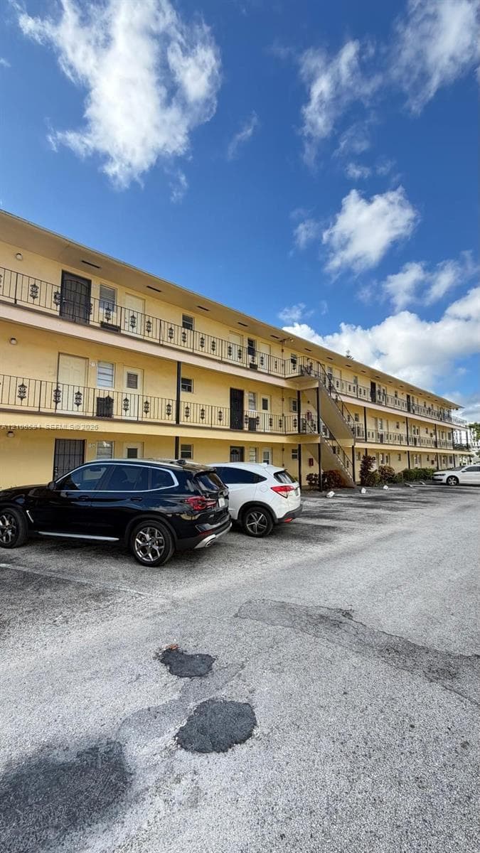Three-story exterior featuring a light-colored stucco facade, decorative black metal railings, external staircases, and dedicated paved parking area