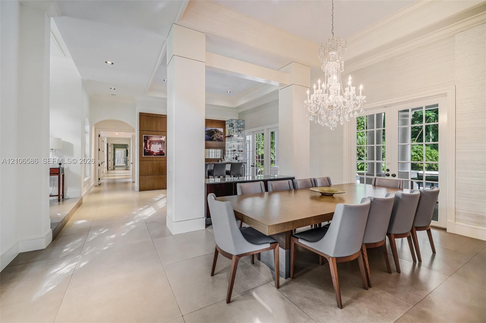 Spacious dining area featuring large-format tile flooring, tray ceilings, floor-to-ceiling columns, a crystal chandelier, and French door access to the exterior