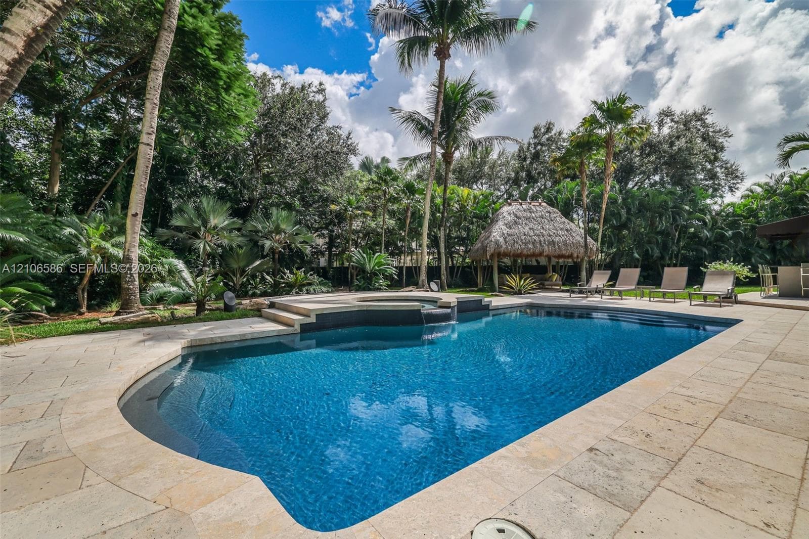 In-ground swimming pool with an integrated elevated spa, stone paver decking, and a detached thatched-roof structure surrounded by mature palms and landscaping