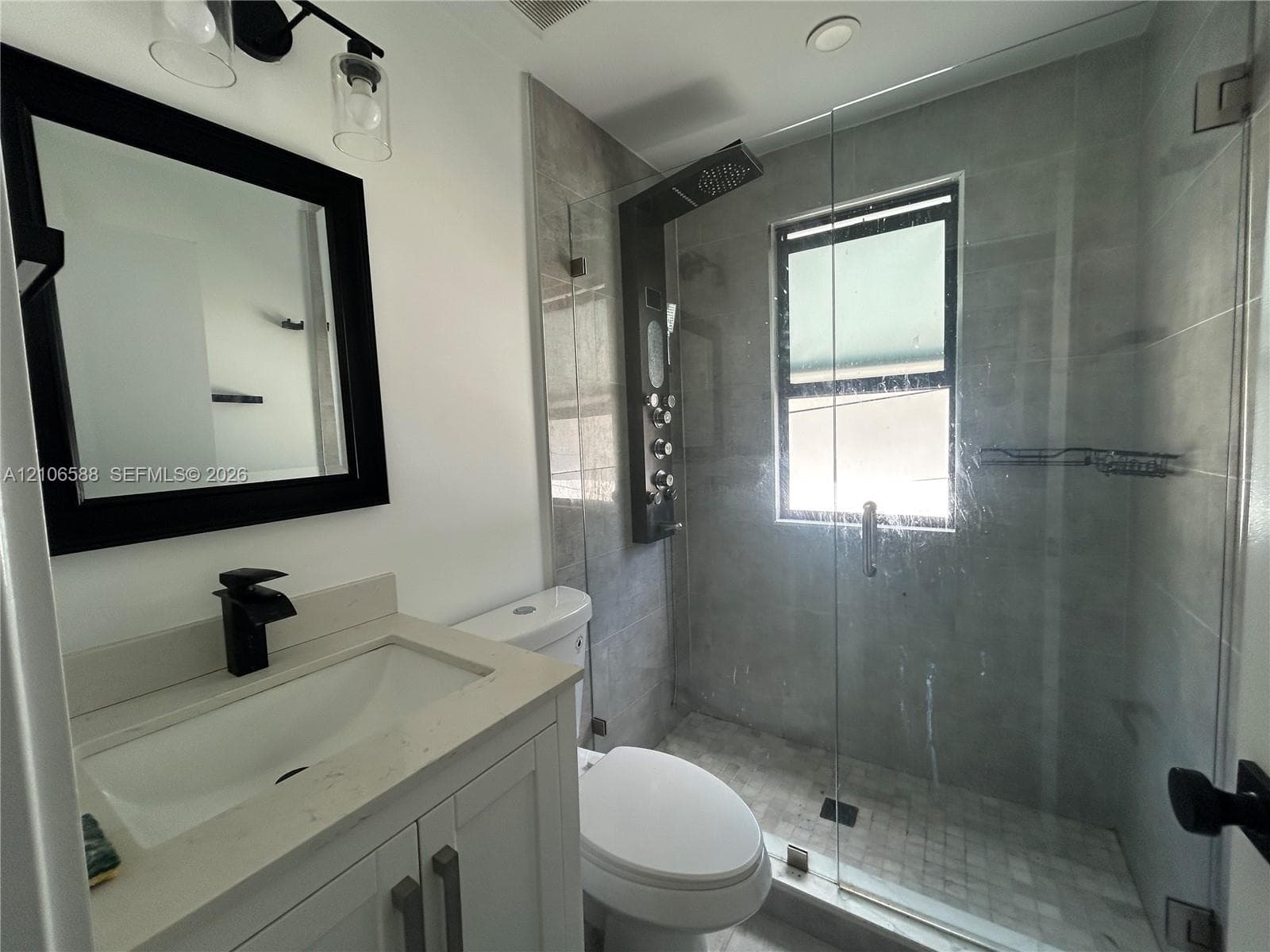 Bathroom featuring a white vanity with a composite countertop, black wall-mounted mirror, matte black faucet, glass-enclosed shower with tiled surround, and a wall-mounted shower panel system