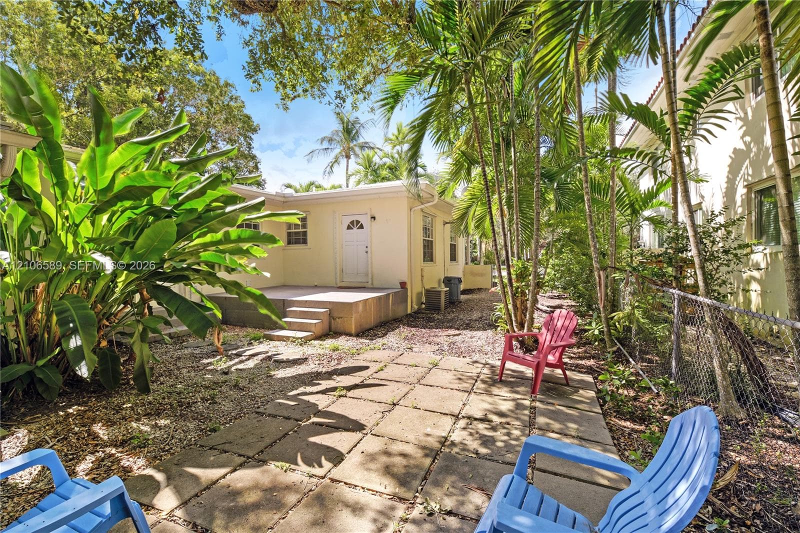 Rear exterior featuring a concrete patio, stone paver ground covering, mature palm trees, and lush tropical greenery